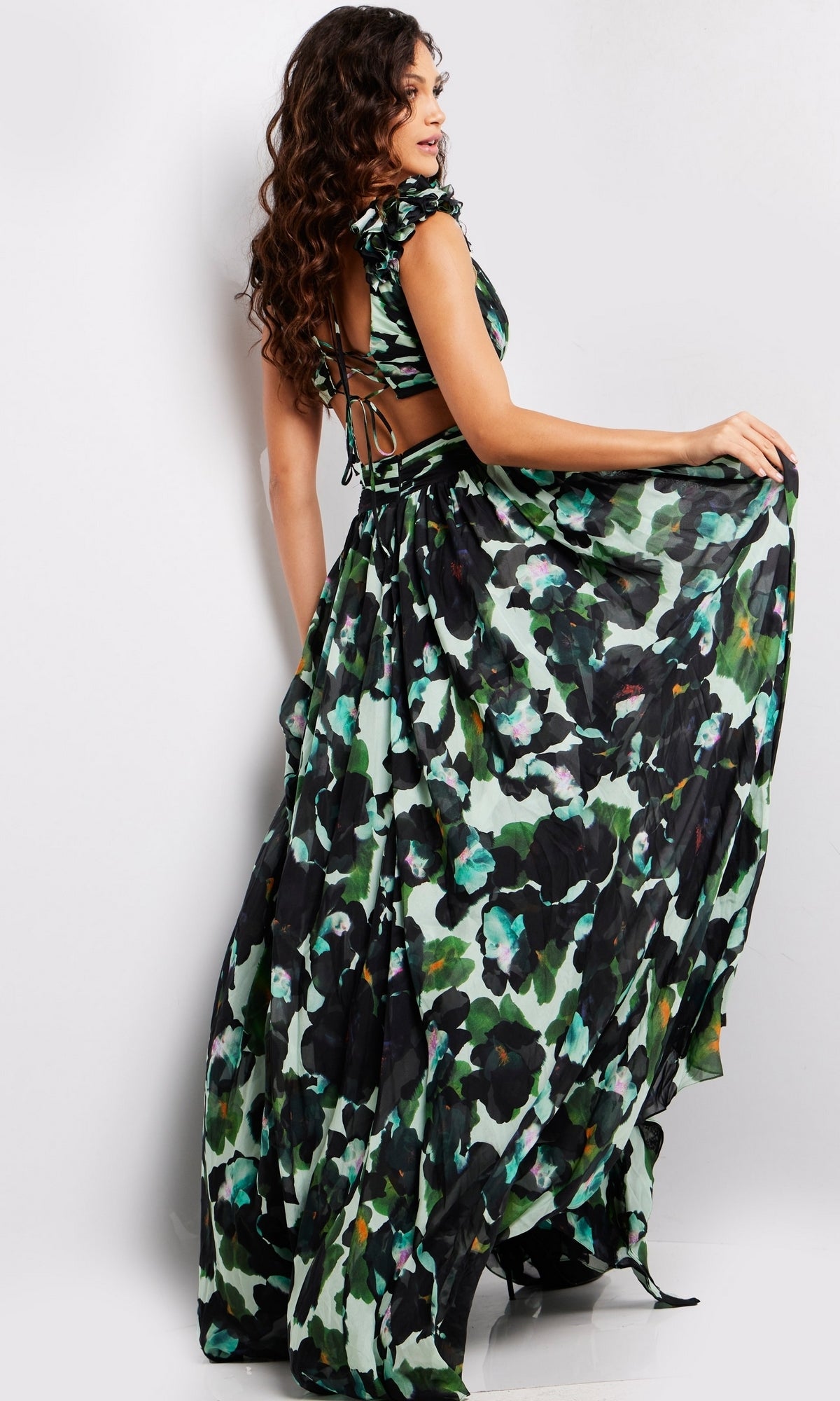 A woman stands holding the skirt of the Long Prom Dress 39420 by Jovani, a flowing green and black floral chiffon gown with an open back and tie details.
